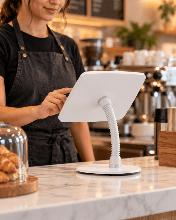 A white modern tabletop monitor with a flexible gooseneck stand and angled screen, placed on a marble counter in a cafe setting, with a female barista in a dark apron interacting with it in the background.